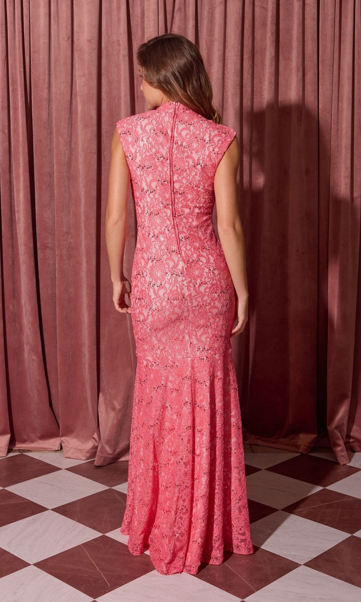 A woman stands on a checkered floor facing away, wearing the Eureka 2061 Long Lace Formal Dress—a sleeveless, floor-length pink lace gown with a high neck—in front of pink curtains.